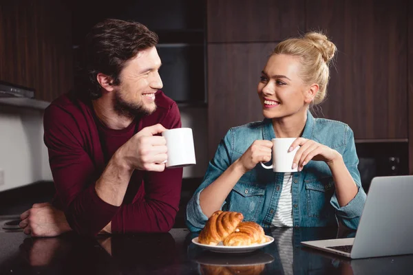 Feliz pareja sosteniendo tazas de café y mirándose en la cocina - foto de stock