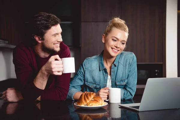 Alegre pareja sosteniendo tazas de café y mirando la pantalla del ordenador portátil en la cocina - foto de stock