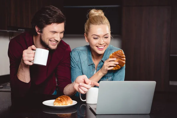 Hombre guapo señalando con el dedo en la pantalla del ordenador portátil mientras chica atractiva sosteniendo croissant en la cocina - foto de stock