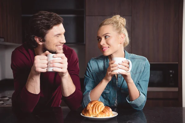 Улыбающаяся пара, держащая чашки кофе и глядя друг на друга на кухне — стоковое фото