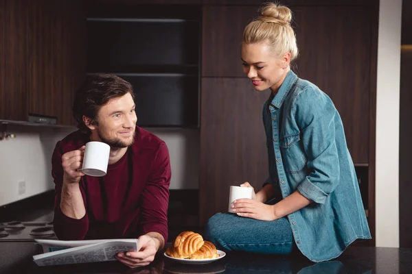 Sonriente pareja sosteniendo tazas de café y leyendo el periódico en la cocina - foto de stock
