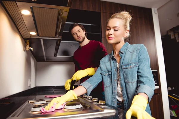 Schöner Mann mit Waschmittelflasche und Blick auf Frau in Gummihandschuhen beim Putzen der Küche — Stockfoto