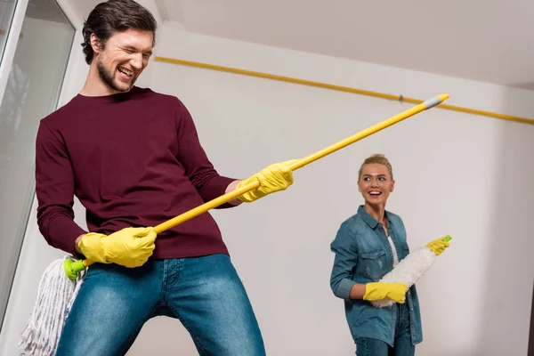 Homem feliz sorrindo e realizando com esfregona e menina alegre segurando espanador no fundo — Fotografia de Stock