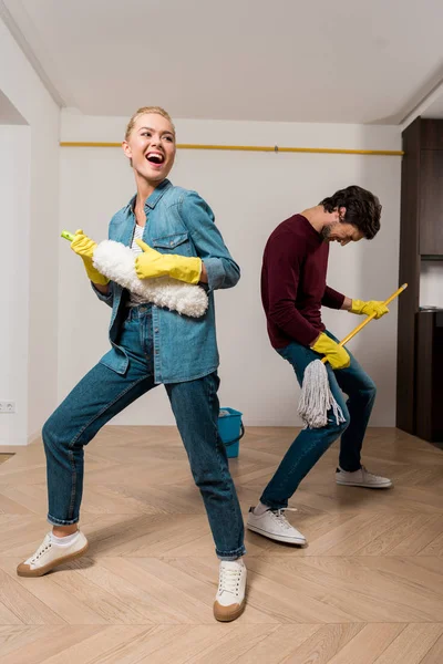 Alegre casal familiar em luvas de borracha marido realizando com esfregona e espanador no apartamento — Fotografia de Stock