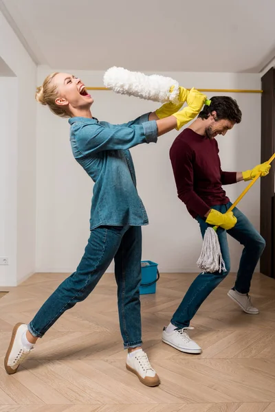 Heureux fille en caoutchouc gants chanter avec poussiéreur tandis que mari effectuer avec la serpillière dans appartement — Photo de stock