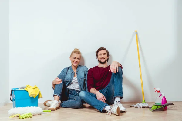 Attractive girl and handsome man sitting on floor near cleaning equipment, looking at camera and smiling in apartment — Stock Photo