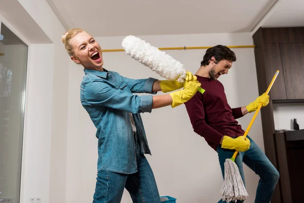 Cheerful girl in rubber gloves singing with duster while husband performing with mop at home — Stock Photo