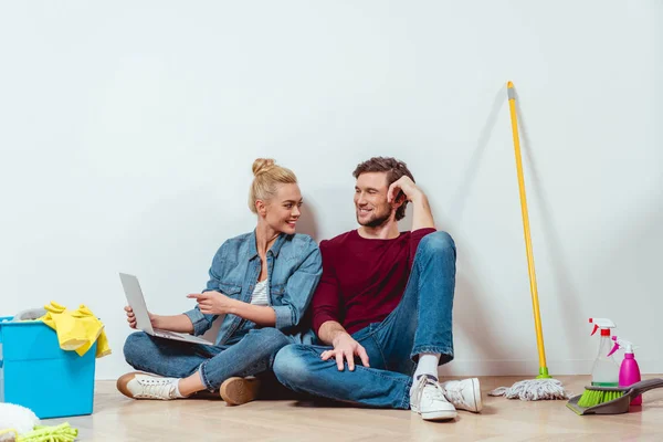 Attraktives Mädchen auf dem Boden sitzend, Ehemann anschauend und mit dem Finger auf Laptop-Bildschirm zeigend — Stockfoto