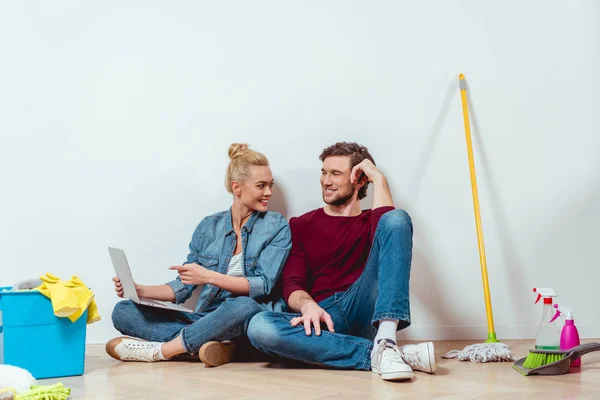 Chica atractiva sentada en el suelo, mirando al marido y señalando con el dedo a la pantalla del ordenador portátil - foto de stock