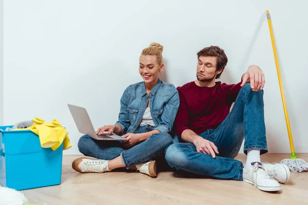 Alegre pareja mirando a la pantalla del ordenador portátil y sentado en el suelo cerca de equipos de limpieza - foto de stock