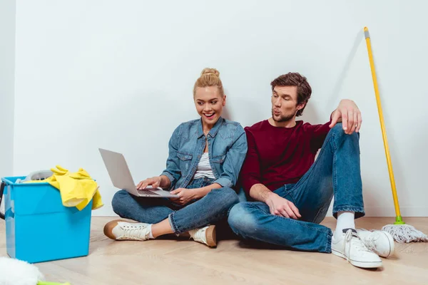 Überraschtes Paar blickt auf Laptop-Bildschirm und sitzt neben Reinigungsgeräten auf dem Boden — Stockfoto
