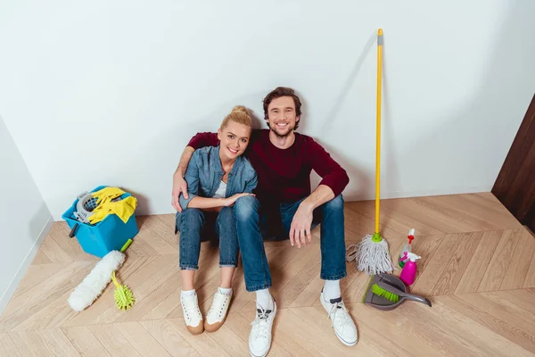 Smiling couple sitting on floor near cleaning equipment and looking at camera — Stock Photo