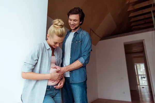 Hombre guapo sonriendo y tocando el vientre de la feliz esposa embarazada en apartamento - foto de stock