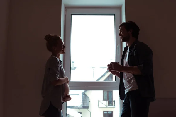 Chica embarazada y hombre guapo de pie junto a la ventana y mirando el uno al otro - foto de stock