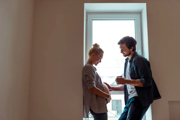 Smiling man standing by window, holding coffee cup and touching belly of pregnant wife — Stock Photo
