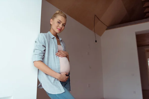 Attractive pregnant girl with hands on belly looking at camera in apartment — Stock Photo