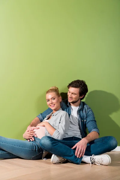 Беременная девушка трогает живот и сидит на полу с улыбающимся мужем за зеленой стеной — стоковое фото