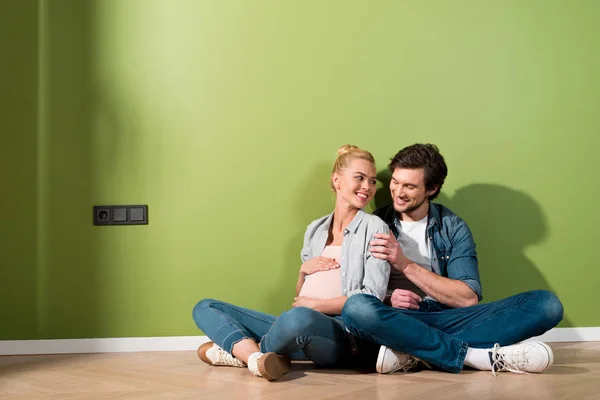 Uomo sorridente seduto sul pavimento e abbracciare moglie incinta — Foto stock
