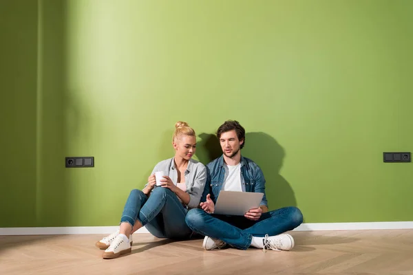 Menina atraente com xícara de café e homem bonito com laptop sentado no chão e falando em apartamento — Fotografia de Stock