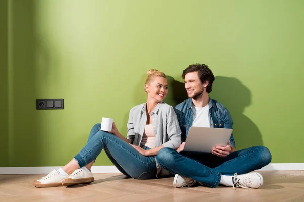 Attractive girl with coffee cup and handsome man with laptop sitting on floor and looking at each other — Stock Photo