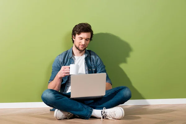 Homem bonito segurando xícara de café, usando laptop e sentado no chão por parede verde — Fotografia de Stock
