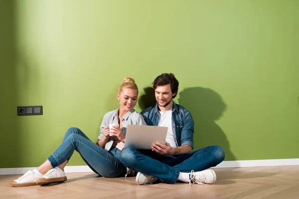 Attractive girl with coffee cup and handsome man sitting on floor and looking at laptop screen — Stock Photo