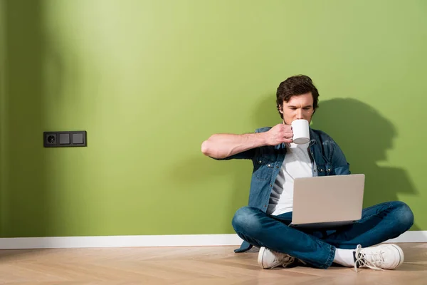 Красивый мужчина пьет кофе, держит ноутбук и сидит на полу у зеленой стены — стоковое фото