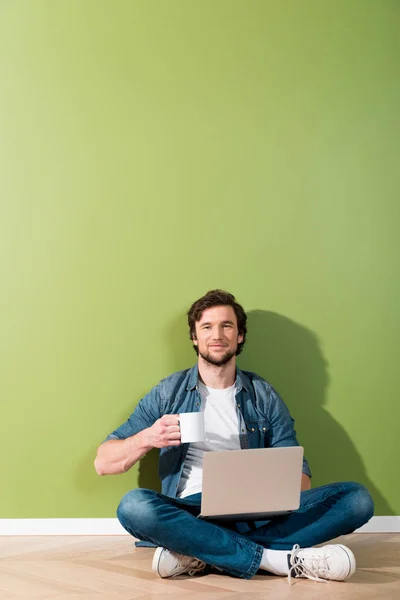 Homme souriant tenant tasse de café et ordinateur portable et assis sur le sol par un mur vert — Photo de stock