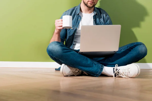 Cropped view of man holding coffee cup and laptop and sitting on floor by green wall — Stock Photo