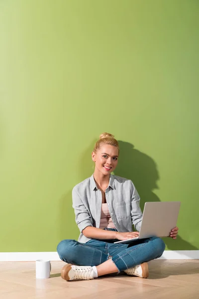 Menina bonita com laptop sentado no chão por parede verde e olhando para a câmera — Fotografia de Stock