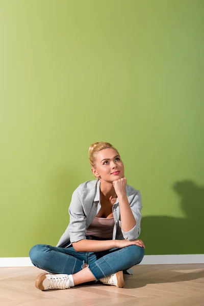 Chica pensativa sentada en el suelo por la pared verde - foto de stock