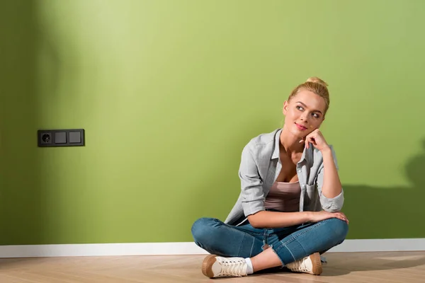Thoughtful girl smiling and sitting on floor by green wall — Stock Photo
