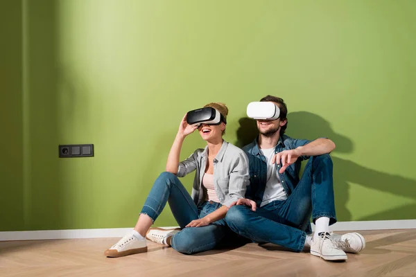 Casal surpreso com fones de ouvido realidade virtual em cabeças sentadas no chão por parede verde — Fotografia de Stock