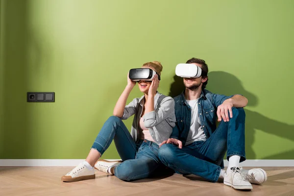 Asombrado chica y hombre con auriculares de realidad virtual en cabezas sentadas en el suelo por la pared verde - foto de stock