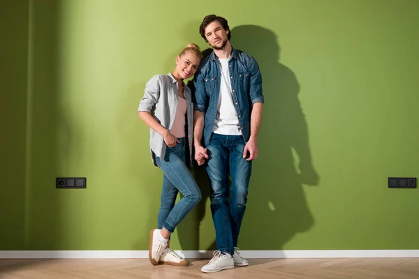 Sonriente pareja cogida de la mano, de pie junto a la pared verde y mirando a la cámara - foto de stock