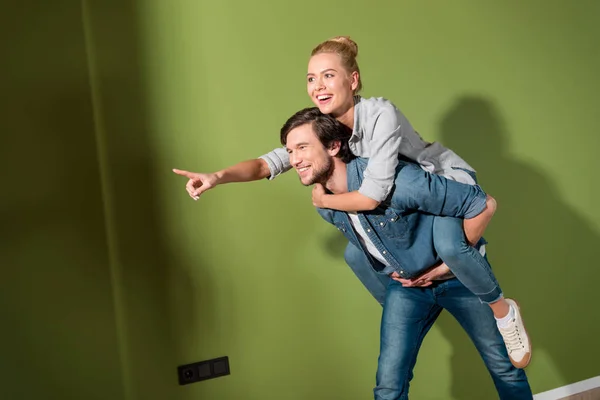 Handsome man giving piggyback ride to beautiful wife pointing with finger in apartment — Stock Photo