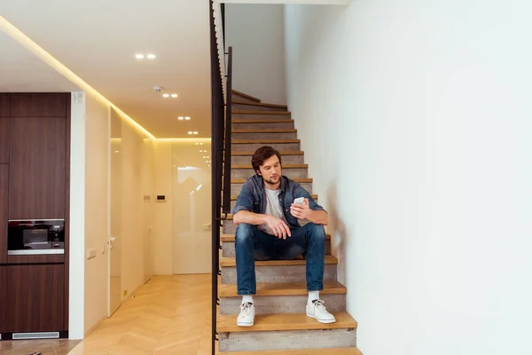 Hombre guapo sentado en las escaleras y mirando la pantalla del teléfono inteligente - foto de stock