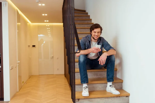 Hombre sonriente sentado en las escaleras y utilizando el teléfono inteligente - foto de stock