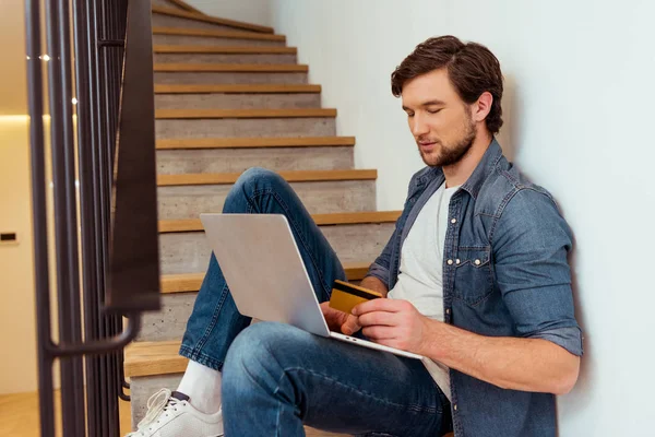 Красивий чоловік сидить на сходах, тримає кредитну картку і використовує ноутбук — стокове фото