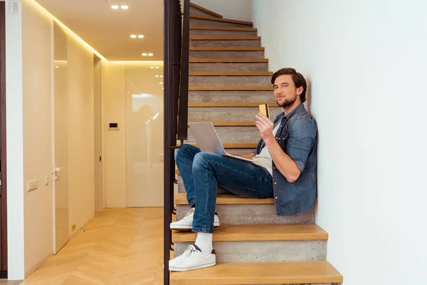 Hombre sonriente con tarjeta de crédito y portátil sentado en las escaleras y mirando a la cámara - foto de stock