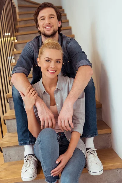 Улыбающийся мужчина, сидящий на лестнице и держащий руки на плечах красивой жены — стоковое фото