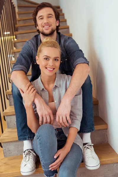 Hombre sonriente sentado en las escaleras y cogido de la mano sobre los hombros de la hermosa esposa - foto de stock