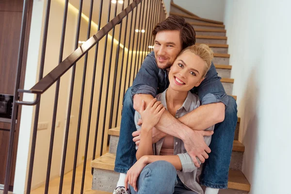 Handsome man sitting on stairs, smiling and embracing beautiful wife — Stock Photo