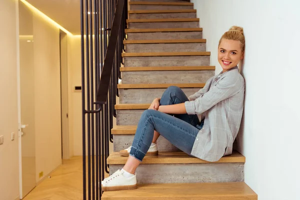Красивая молодая девушка сидит на лестнице, улыбается и смотрит в камеру — стоковое фото