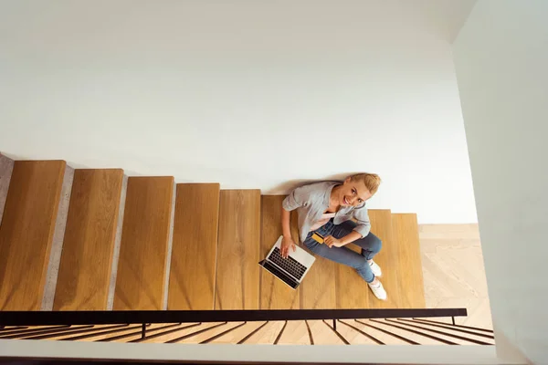 Blick von oben auf lächelndes Mädchen, das mit Laptop auf der Treppe sitzt und in die Kamera schaut — Stockfoto