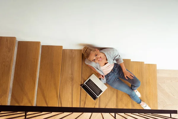 Vista aérea de menina atraente sentado com laptop nas escadas e olhando para a câmera — Fotografia de Stock