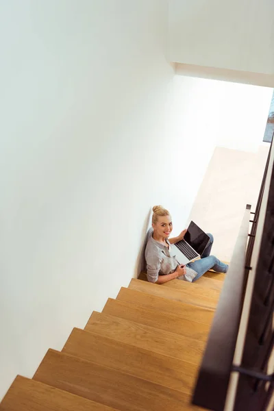 Belle fille assise sur les escaliers, en utilisant un ordinateur portable et en regardant la caméra — Photo de stock