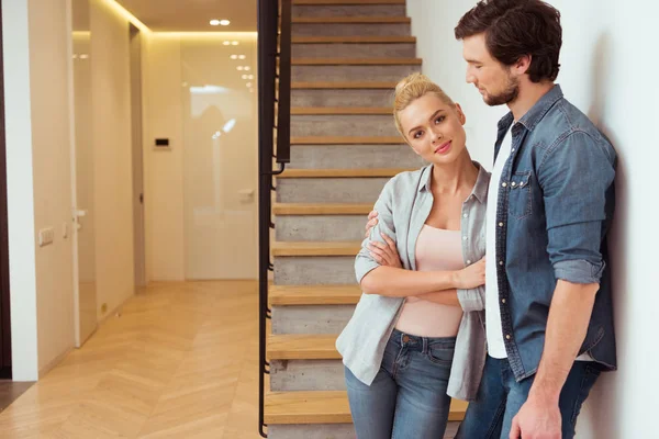 Menina atraente olhando para a câmera e de pé com o marido perto de escadas em casa — Fotografia de Stock