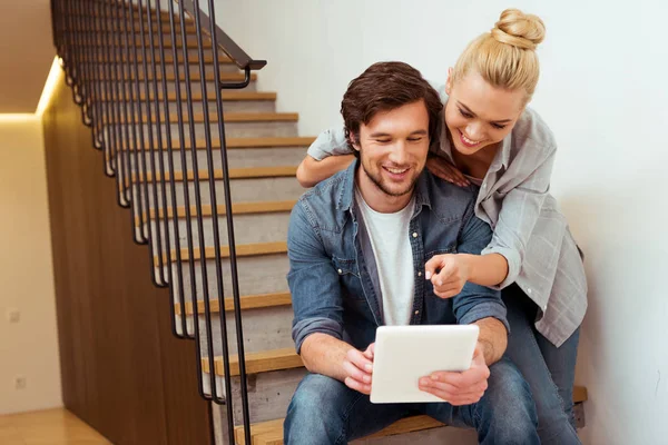 Handsome man sitting on stairs and holding digital tablet with wife pointing with finger at screen — Stock Photo