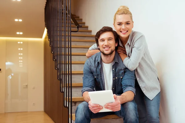 Handsome man sitting on stairs and holding digital tablet with smiling wife at home — Stock Photo